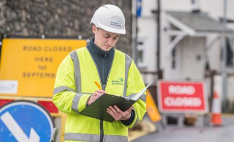 A Northern Gas Networks employee wearing a hi-vis vest and safety headwear, writing in a notebook. Various roadside construction equipment is in the background.