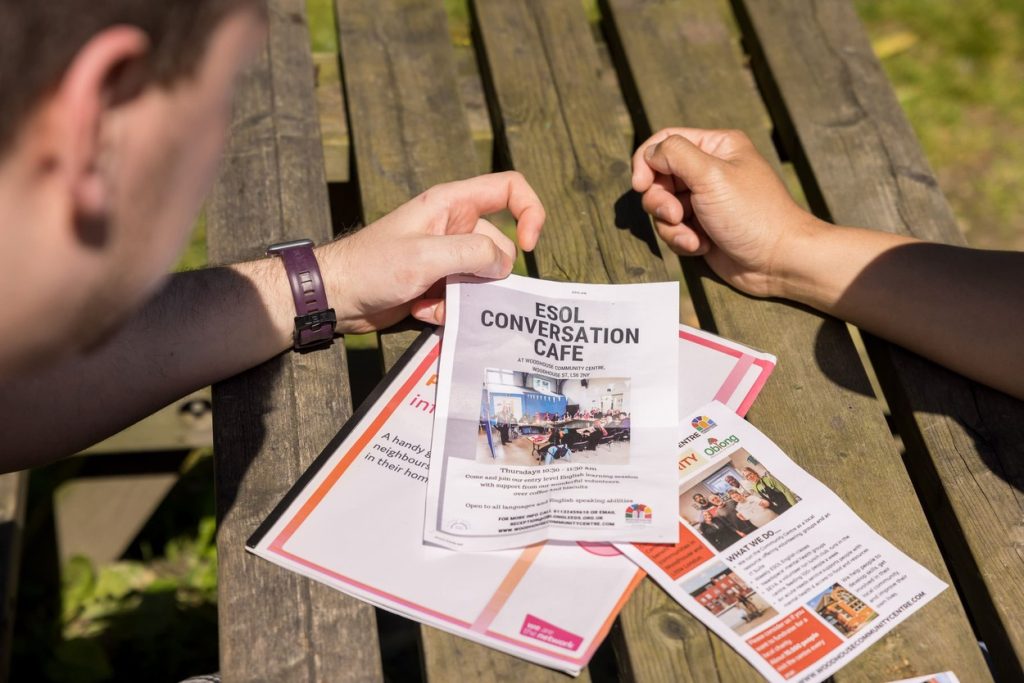Image of two people looking at a series of flyers/posters on a table outside. The foremost flier reads "ESOL Conversation Cafe".