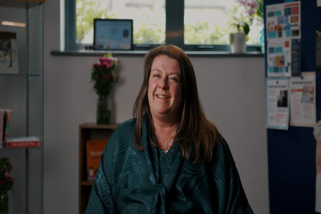 Image of a smiling lady with brown hair in a green top. A glass cabinet housing books, a small bookshelf with a vase of flowers, a window and a mounting board with various posters and infosheets are in the background, slightly blurred.