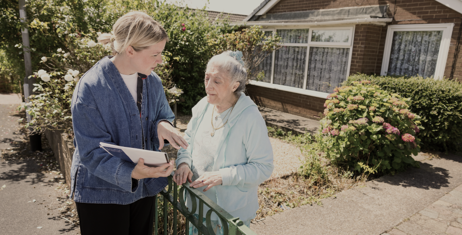 Image of a lady in a blue shirt showing some documents to an elderly lady outside a house